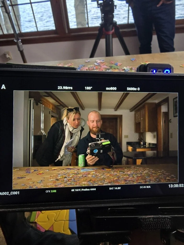 Two people working with a puzzle on a table, with a camera capturing their photo. One person is holding a camera, and the other person is leaning in, both looking at the camera. The scene is indoors with wooden cabinets in the background.