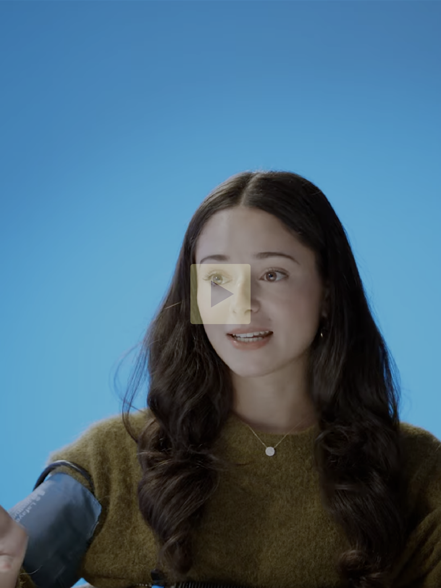 A young woman with long dark hair, wearing a brown sweater and a delicate necklace, sitting against a blue background, talking.