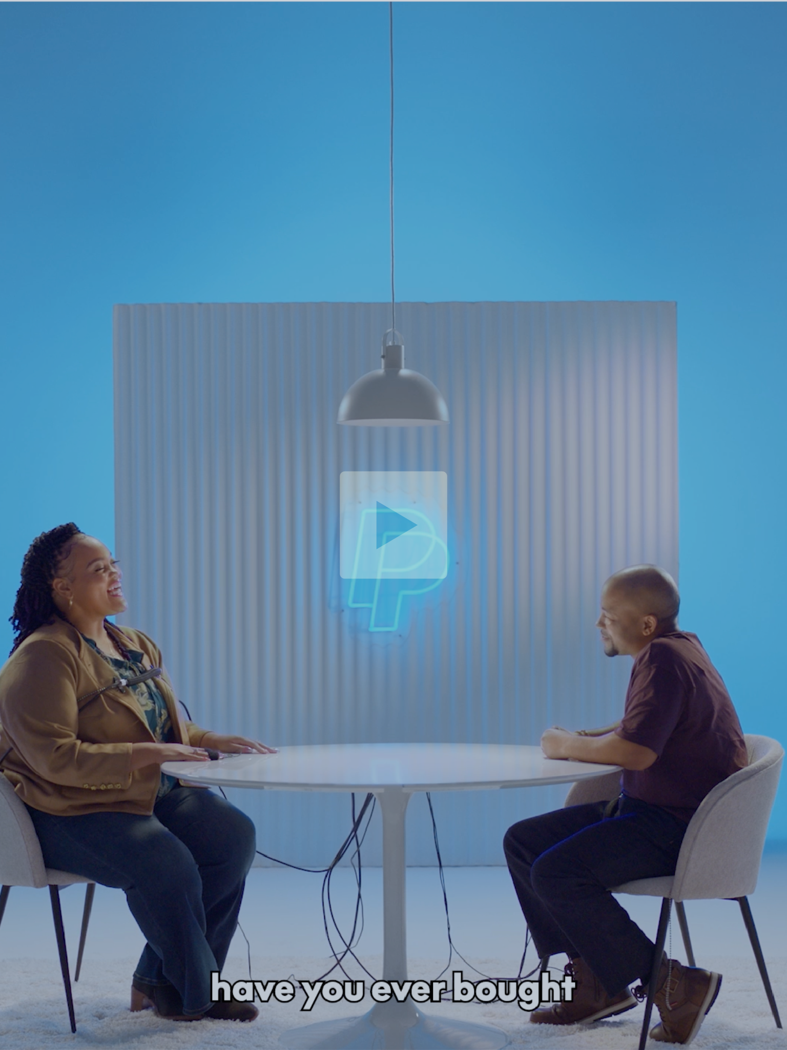 Two people sitting at a round table, chatting and laughing, in a brightly lit room with a blue background and a neon '4' sign.
