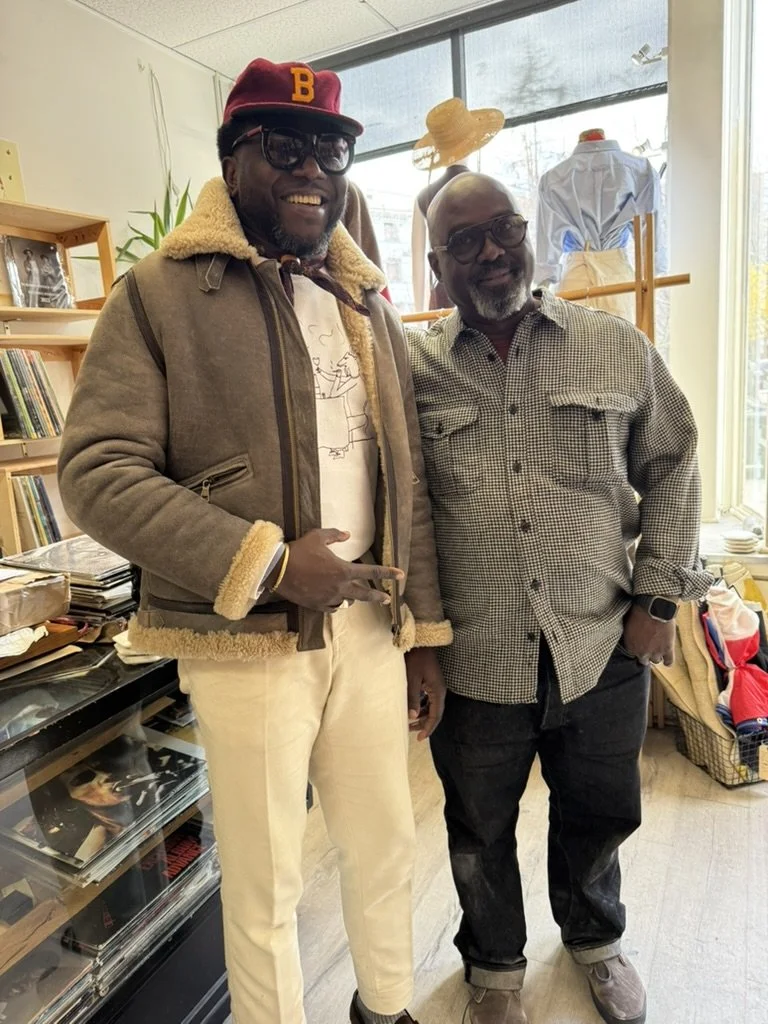 Two men with dark skin standing inside a store or library, smiling at the camera. The man on the left wears a brown leather jacket with a shearling collar, cream-colored pants, a red hat with a yellow 'B,' sunglasses, and a white t-shirt. The man on 