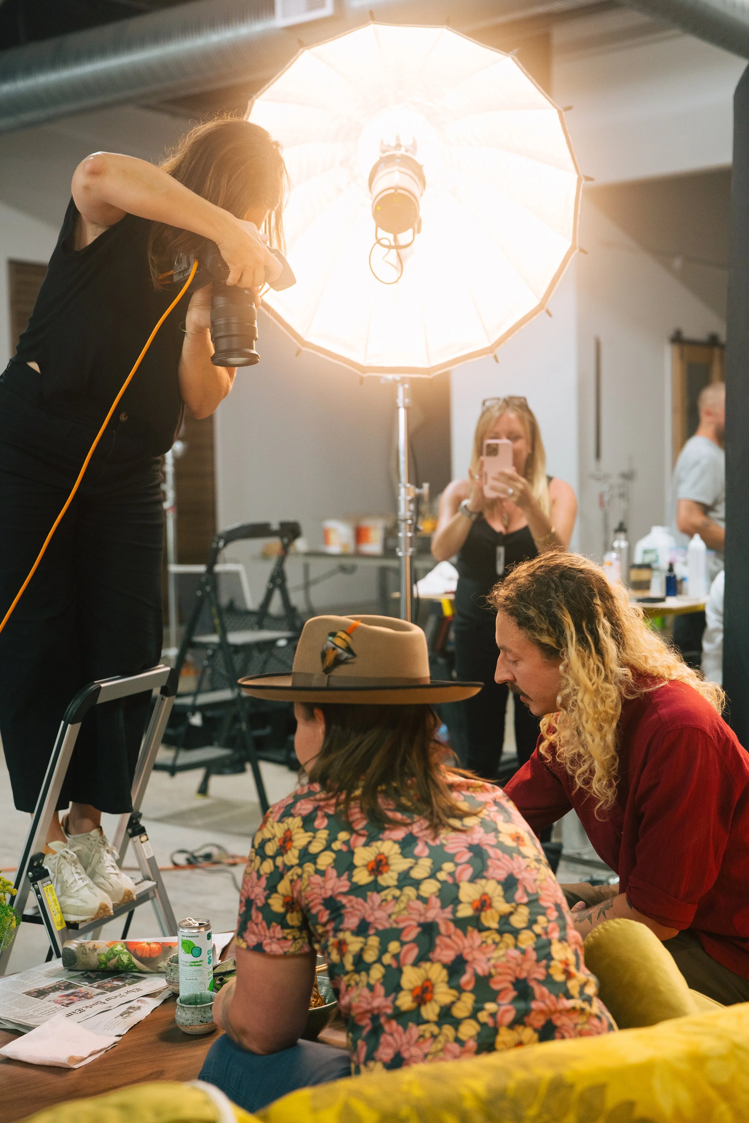 Behind the scenes of a photoshoot or film set with a large umbrella lighting, a woman on a ladder adjusting camera, another woman taking a photo with her phone, and two people sitting and talking at a table surrounded by various items and supplies.