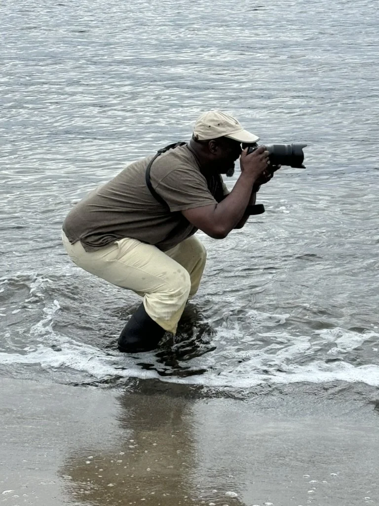 A person standing in shallow water at the beach, holding a camera and taking photos.