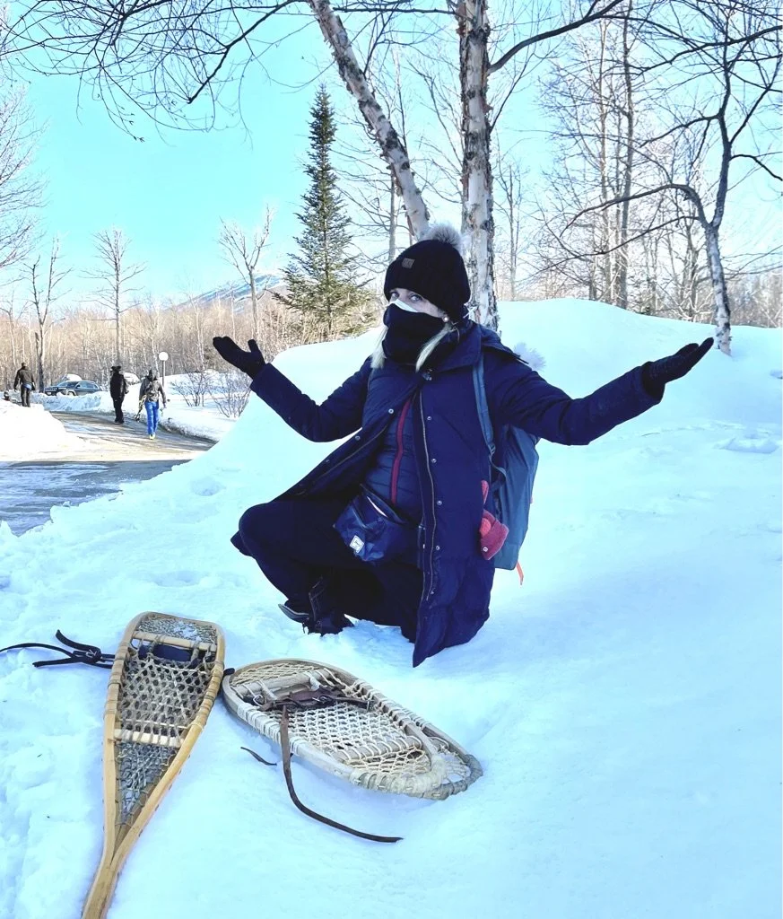 Person kneeling in snow with snowshoes nearby, outdoors in winter, wearing a black coat, black hat, face mask, and gloves, with a backpack, gesturing with arms outstretched, surrounded by snow-covered trees and other people in the background.