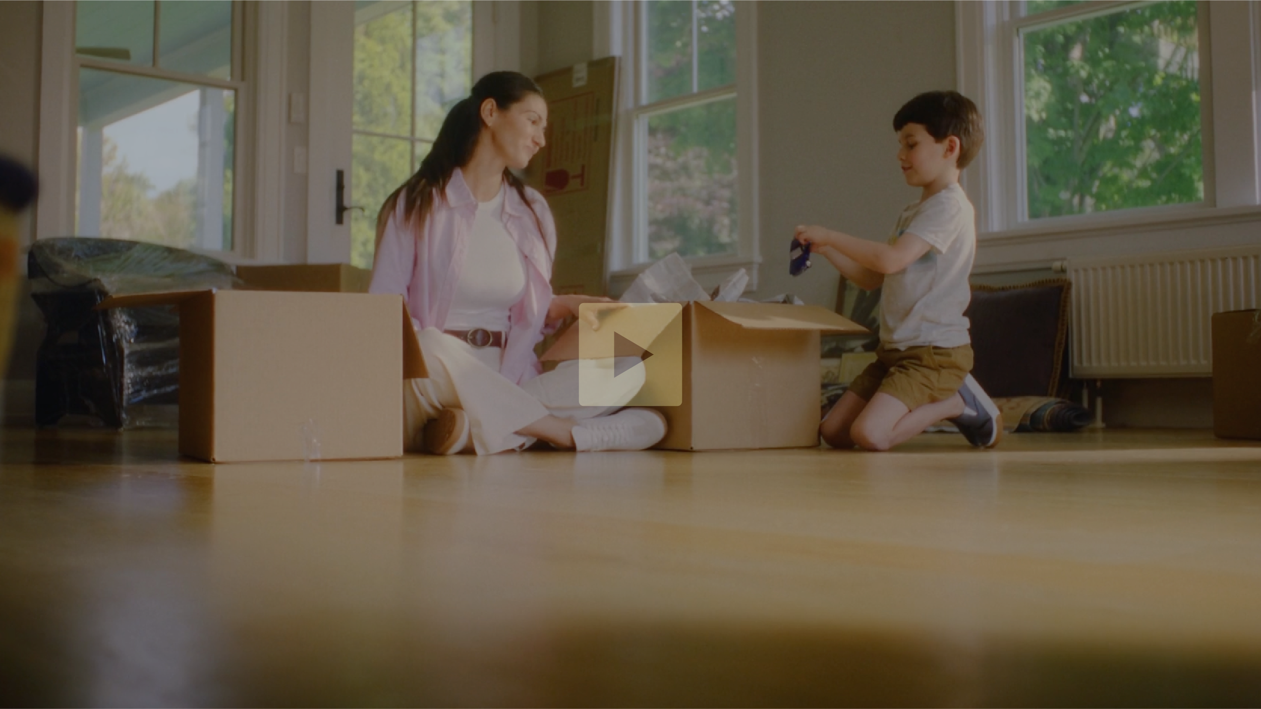 A woman and a young boy sit on the floor among moving boxes in a sunlit room, with large windows showing green trees outside, as they pack or unpack belongings.