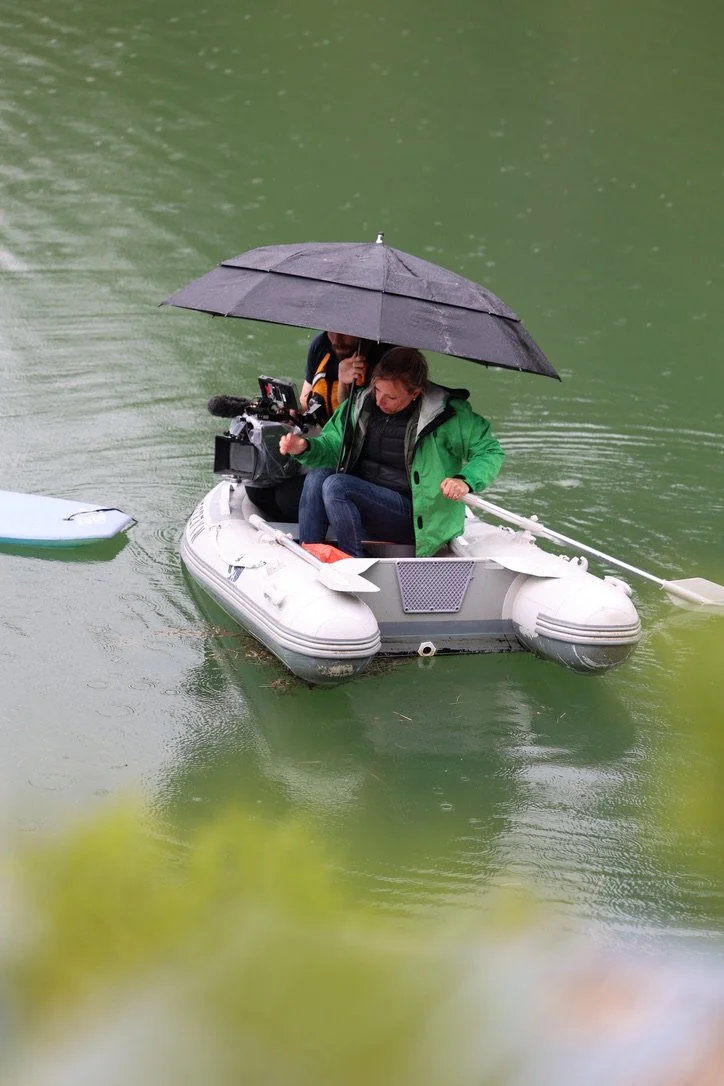 Person sitting in an inflatable boat holding an umbrella, with camera equipment nearby on green water.