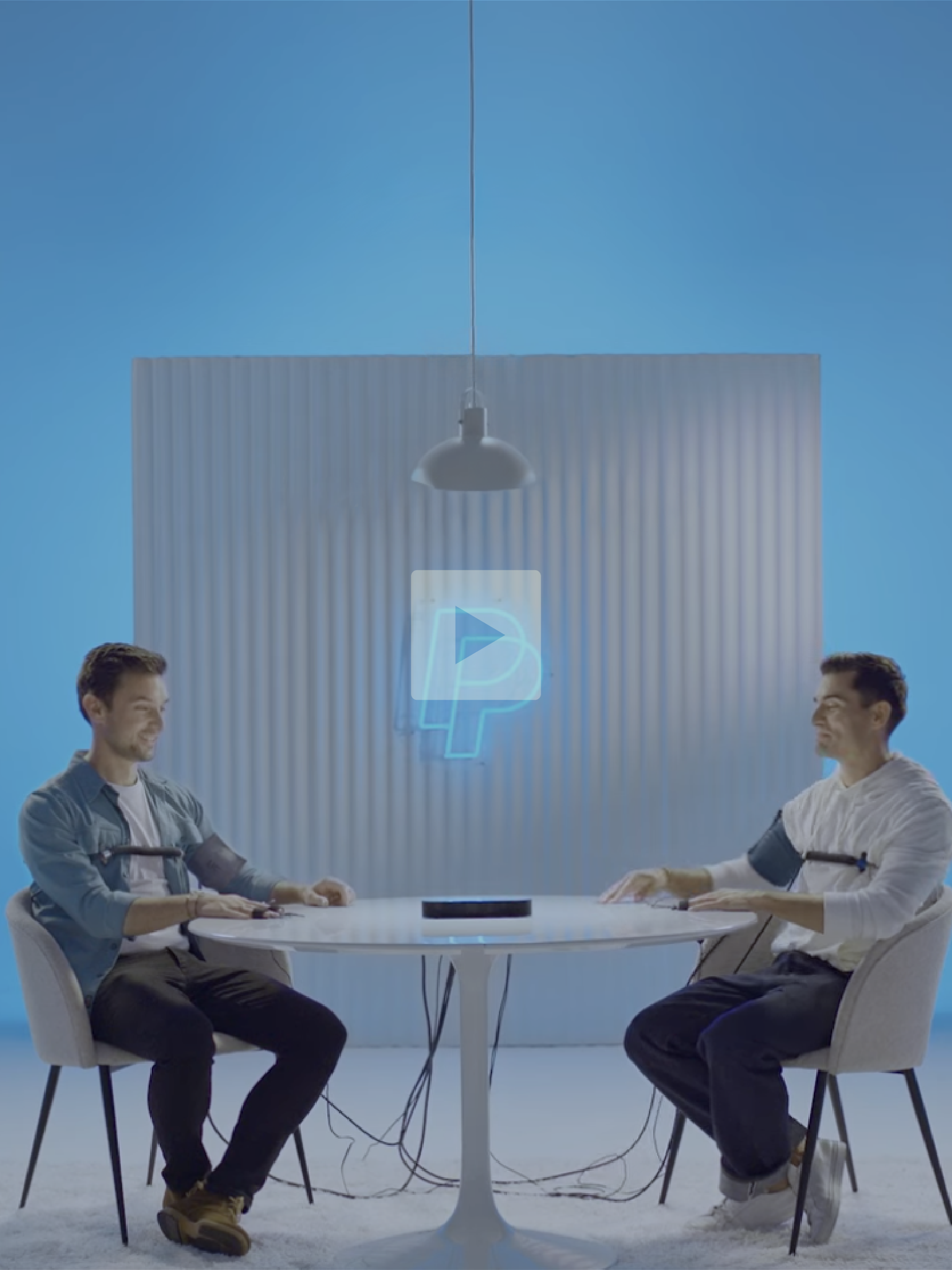 Two young men sit at a round table facing each other, playing a video game with wired controllers. They are smiling and appear to be enjoying the game. A neon blue '4' logo is projected on the wall behind them, which features a blue background and a white vertical panel with a hanging light fixture above. The scene suggests a modern, playful gaming setup.