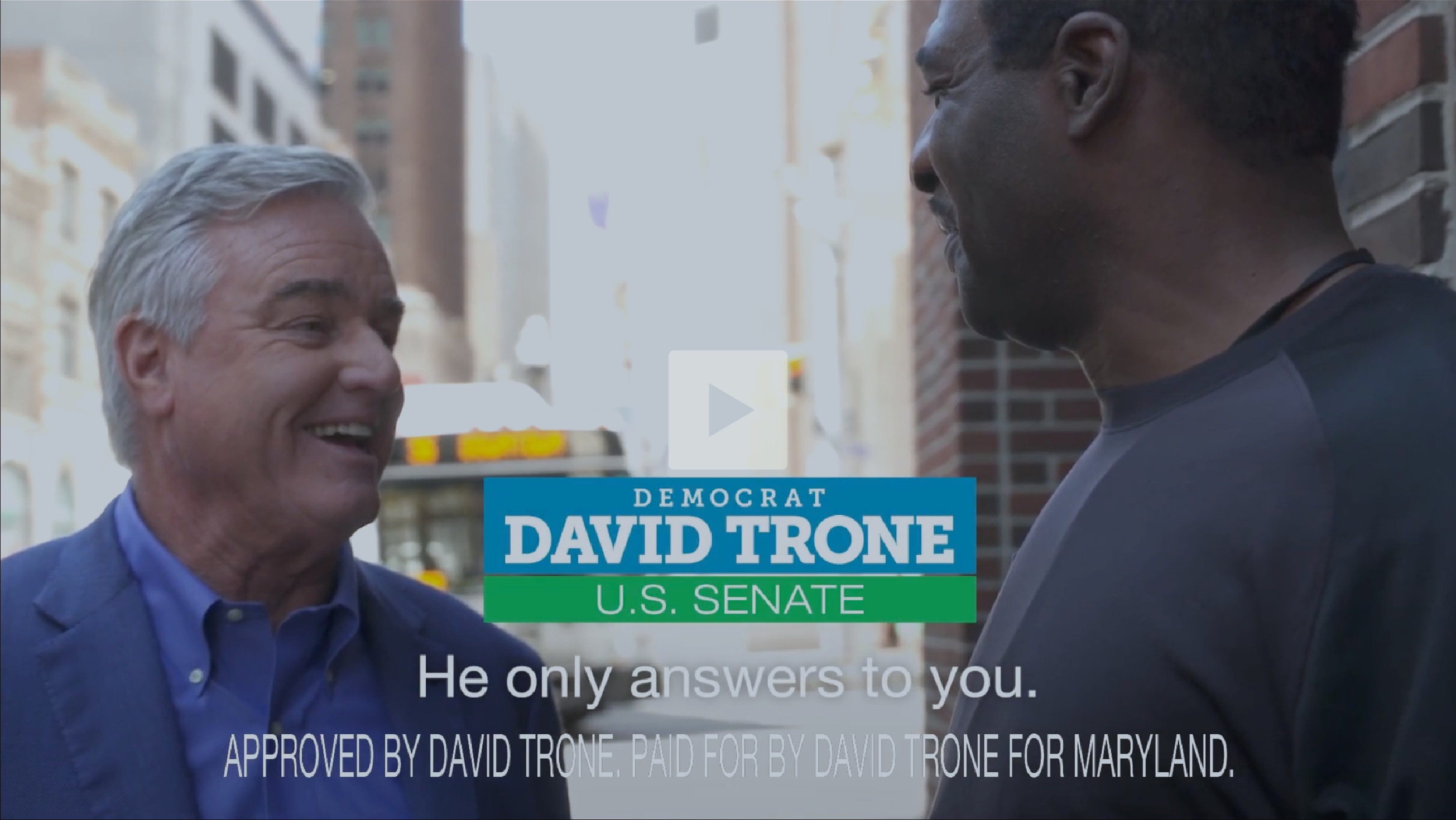 Screenshot of two men standing on a city street, engaged in conversation. One man has gray hair and is wearing a blue blazer, smiling. The other man has dark hair and is wearing a black shirt, facing him. There is an overlay of campaign text for David Trone, a U.S. Senate candidate, including his name and district, with some subtitles.