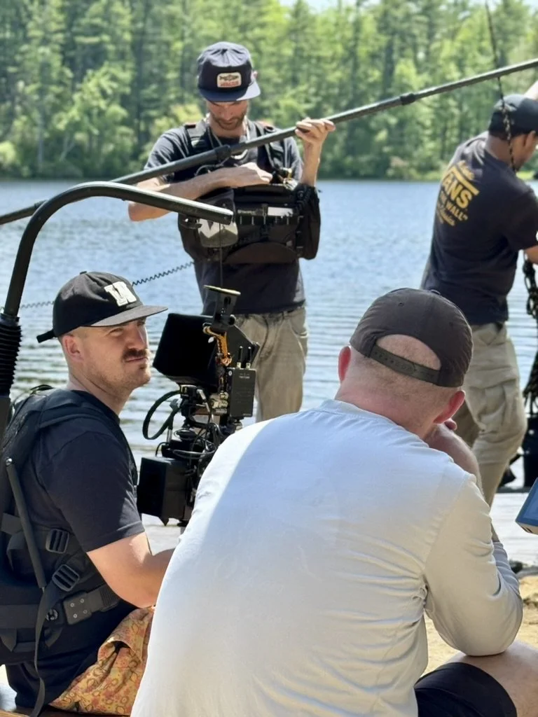 A film crew is filming at a lakeside. One person is operating a camera, while another is holding a boom microphone. Two other crew members are sitting in front, one wearing a cap and the other with a backwards cap, possibly engaging in a discussion or observing the scene.