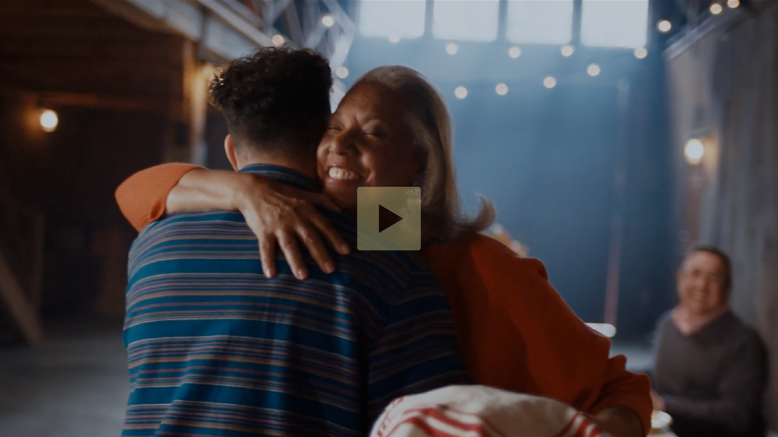 Two people hugging warmly, smiling with joy, in a warmly lit indoor setting with string lights and wooden walls, while a third person in the background also smiles.