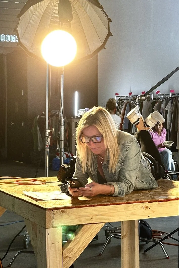 A woman with blonde hair and glasses laying on her stomach on a wooden table, looking at her smartphone, in a room with clothing racks and people in the background, illuminated by a large professional light.