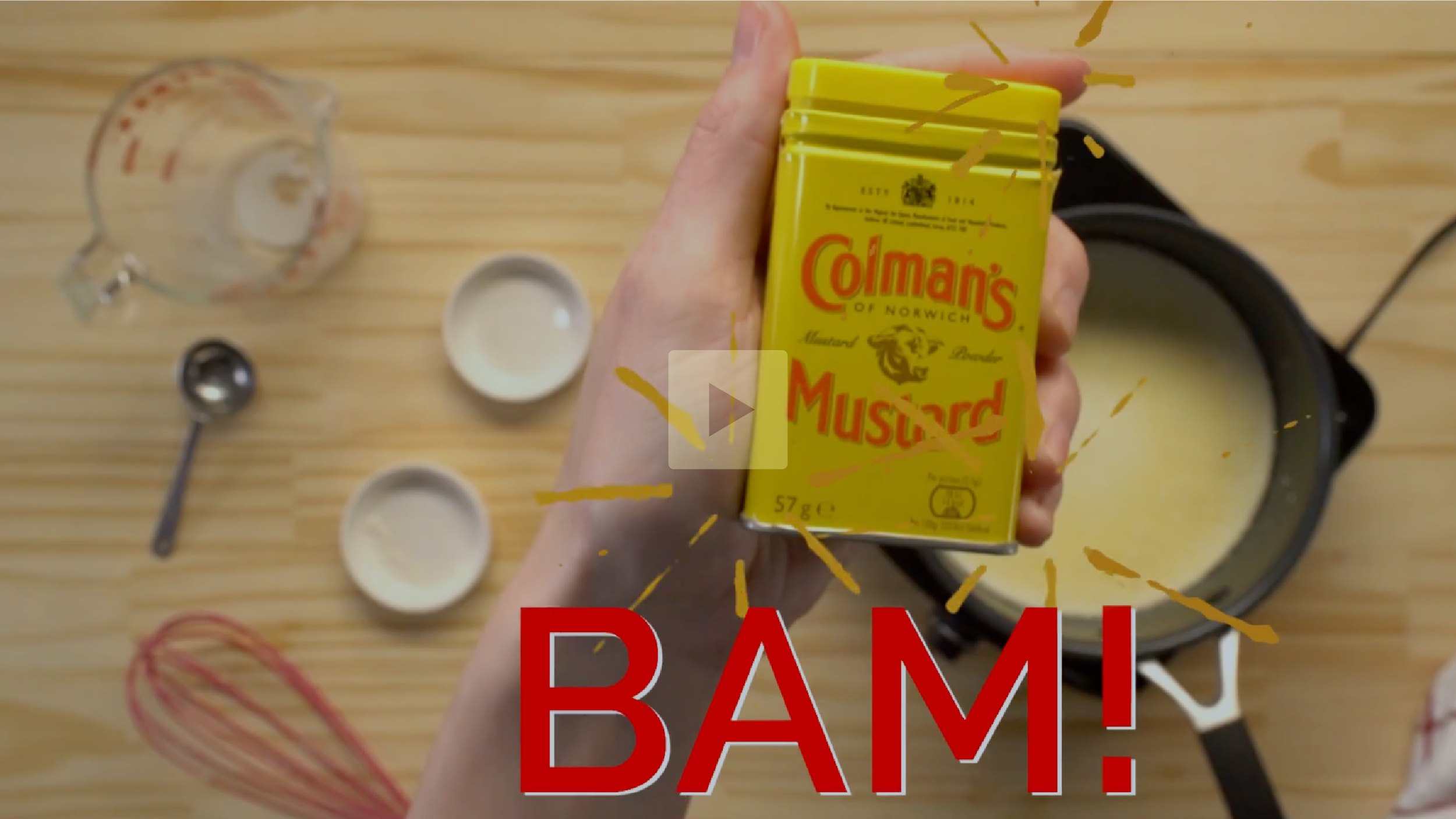 A hand holding a yellow box of Colman's mustard over a wooden countertop with measuring cups, a whisk, and a pot with cream or sauce.