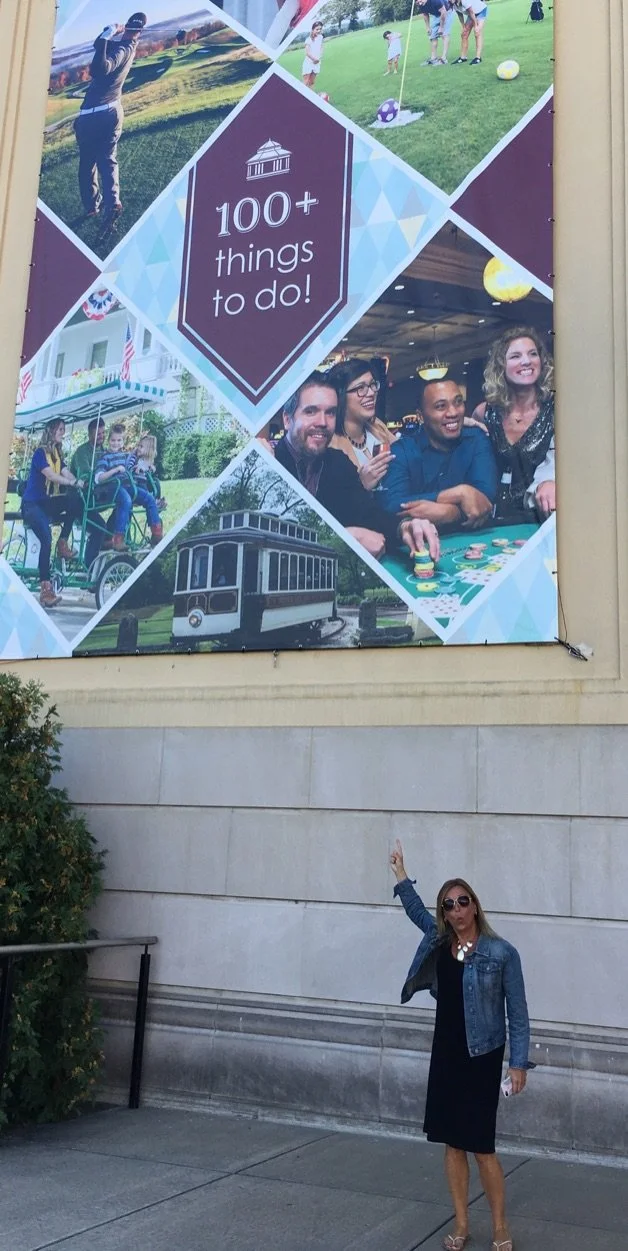 A woman in sunglasses and a denim jacket pointing at a large outdoor billboard featuring various images of people engaging in outdoor activities, including playing soccer, riding a bicycle, and socializing at a casino, with the text '100+ things to d