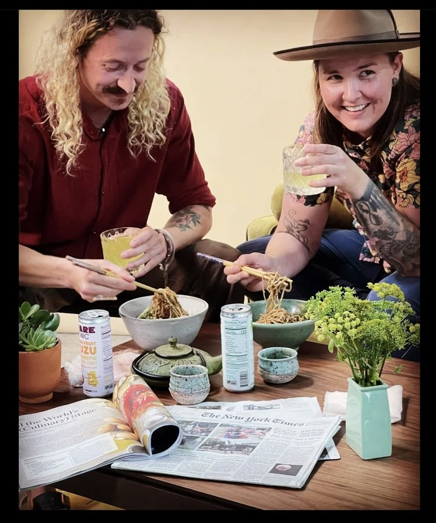 Two women sit at a table eating noodles. One woman has curly blonde hair, a mustache, and wears a red shirt. The other woman has straight hair, tattoos on her arms, and wears a wide-brimmed hat and a floral shirt. They are smiling and holding glasses