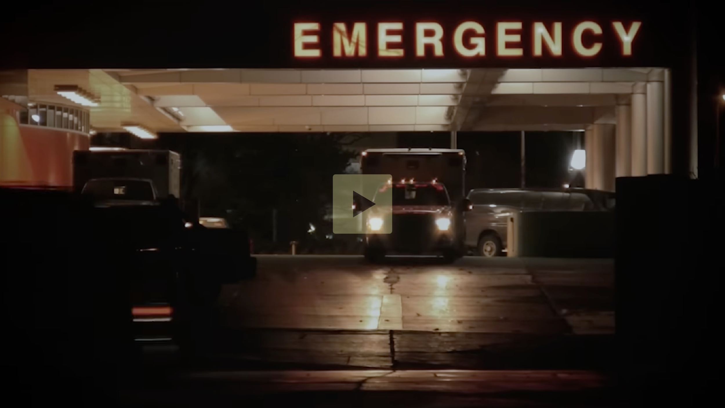 Nighttime view of an emergency entrance with a large illuminated "EMERGENCY" sign, vehicles including an ambulance entering, and a dark exterior setting.
