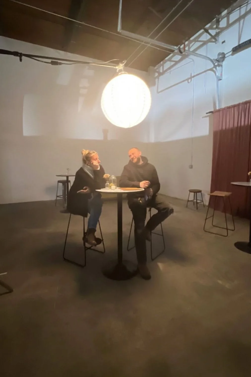 Two people sitting at a high round table in a minimalist indoor space, smiling and talking, with a large bright circular light overhead and bare stools in the background.