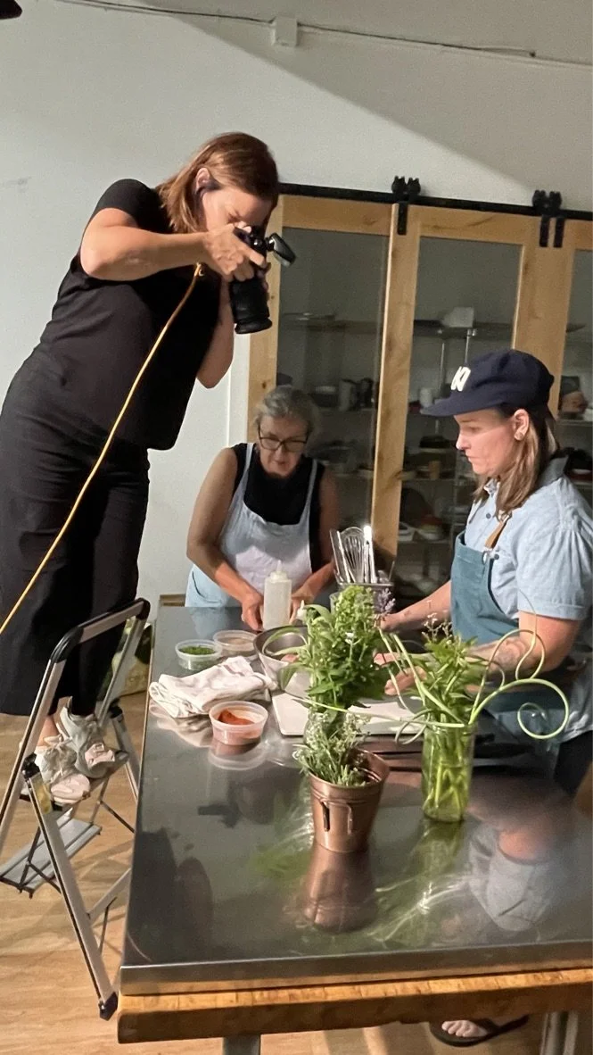 Two women preparing food in a kitchen with a photographer capturing the moment. One woman is wearing an apron and measuring ingredients, while the other is standing on a step stool taking photos. There are potted herbs, bowls, and kitchen utensils on