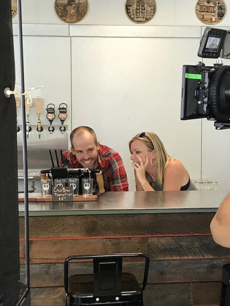 Two people, a man and a woman, are sitting at a bar counter, laughing and enjoying a tasting flight of drinks. There is a camera on a tripod capturing the moment.