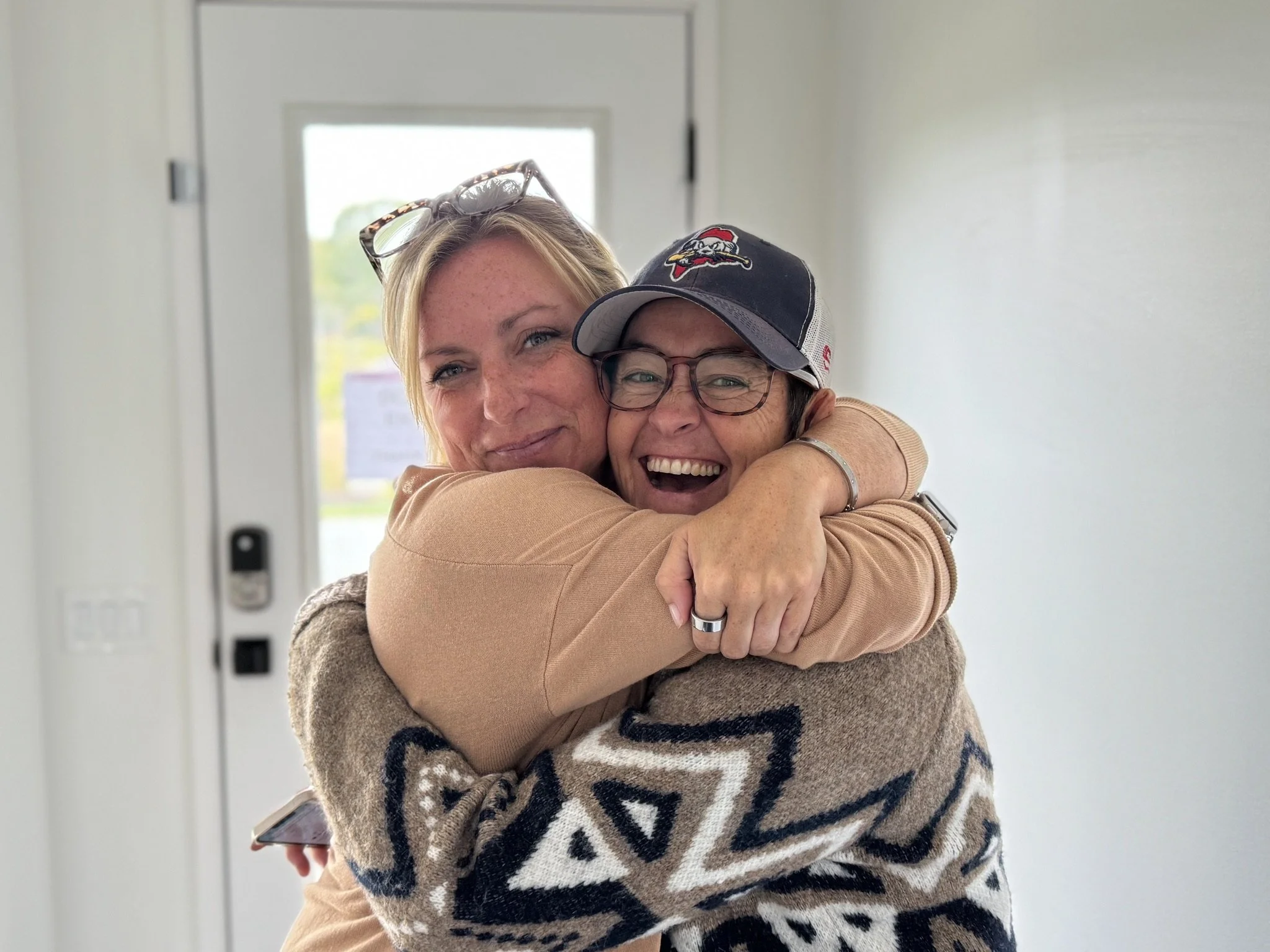 Two women hugging and smiling inside a house, with one woman wearing glasses and a cap with a sports team logo, and the other woman wearing glasses and a patterned sweater.