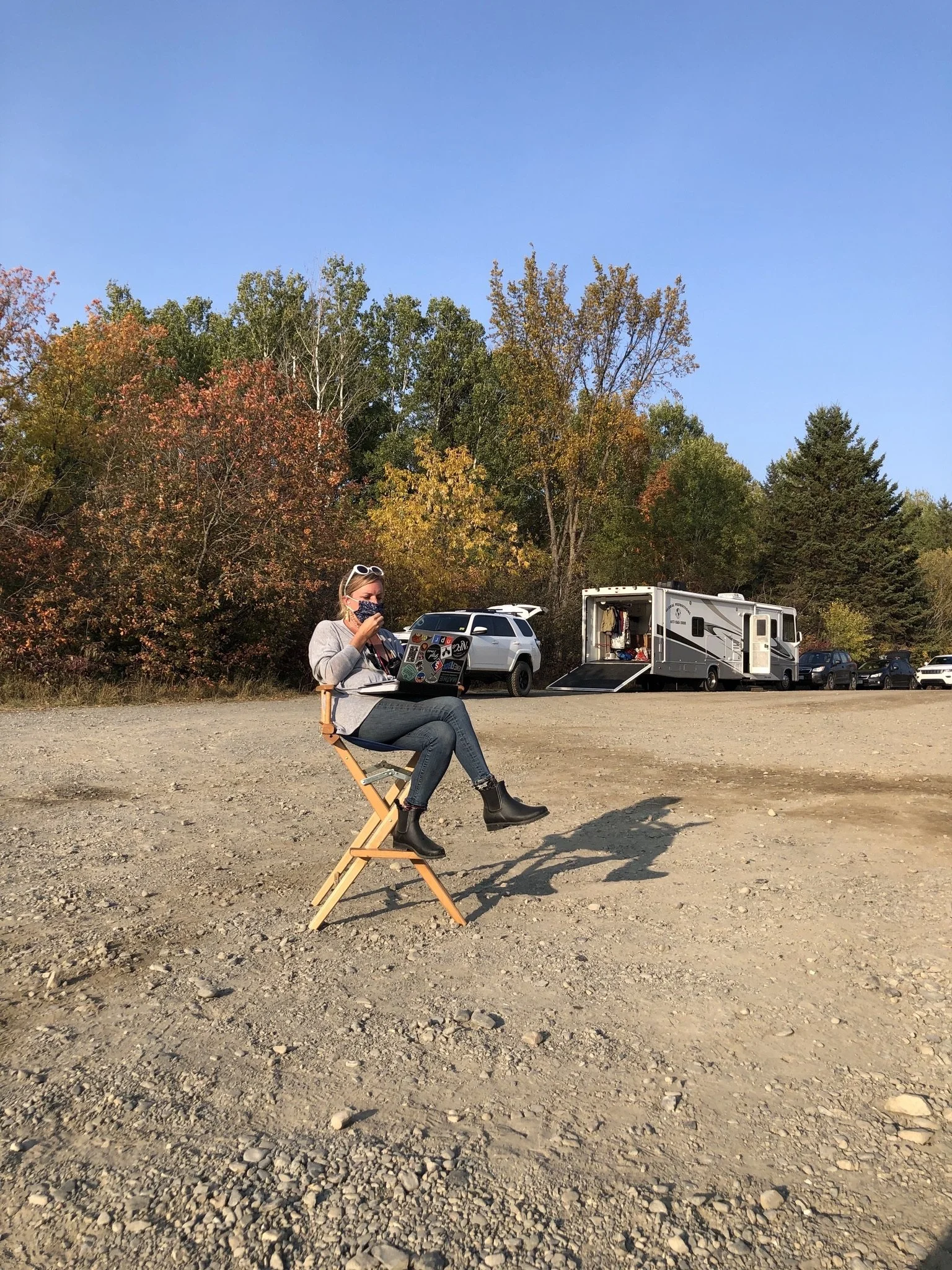 A woman sitting on a folding chair outdoors, working on a laptop in a gravel area. She is wearing a mask, sunglasses, and boots. In the background, there are several parked cars and a large RV, with trees showing fall foliage under a clear blue sky.