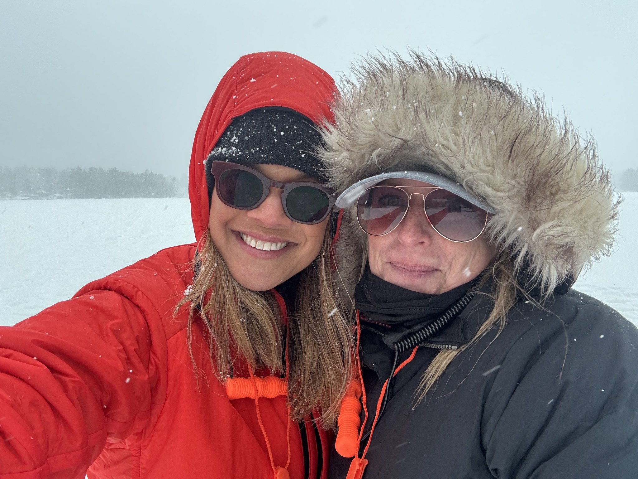 Two women in winter clothing, sunglasses, and hooded jackets smiling outdoors in a snowy landscape.