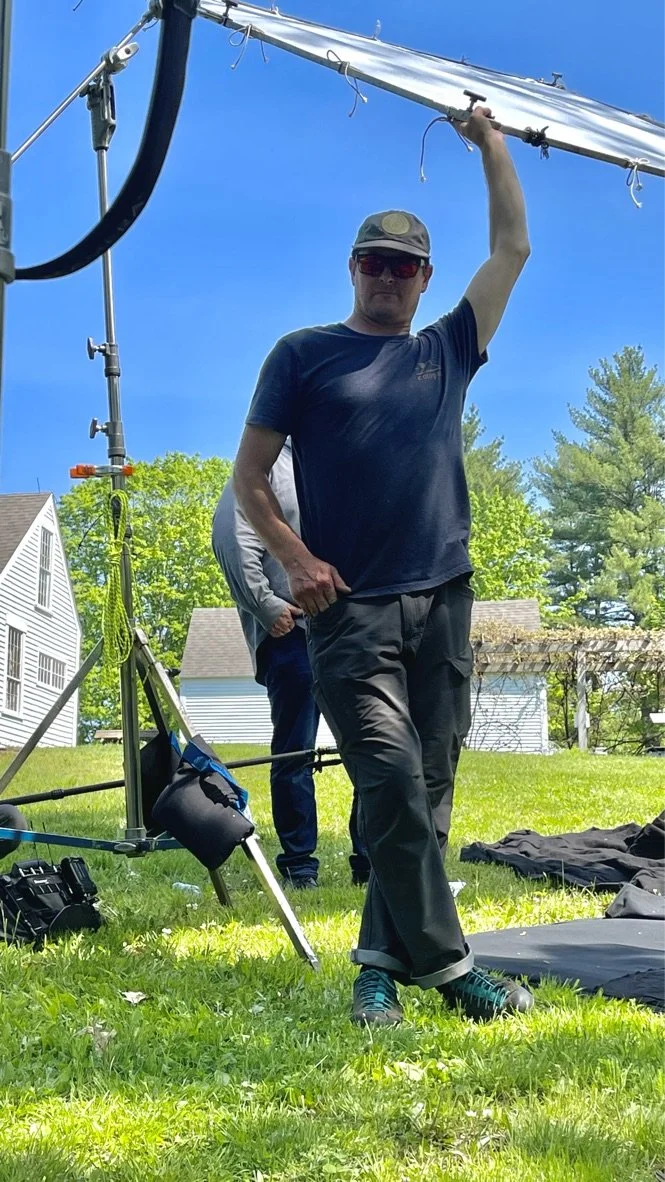 A man operating camera equipment outdoors on a grassy area with trees and houses in the background, weather sunny with clear blue skies.