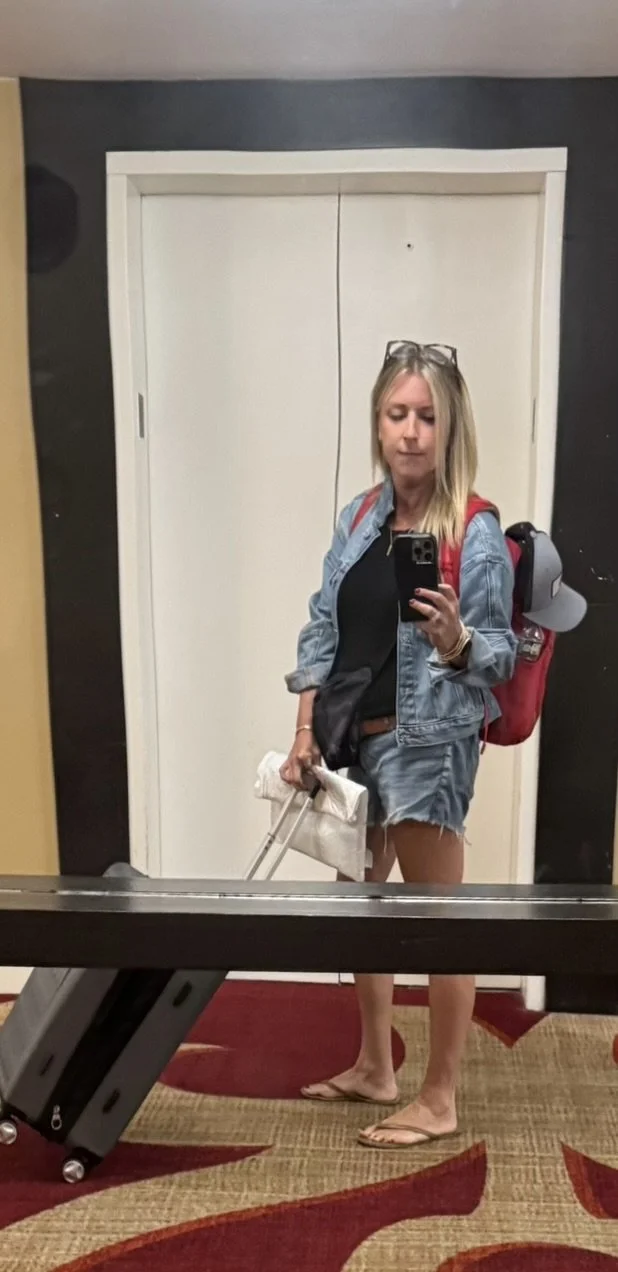 A woman taking a mirror selfie in a hotel hallway, dressed in a denim jacket, black top, jean shorts, flip-flops, with a red backpack and sunglasses on her head, carrying a white bag and pulling a suitcase.