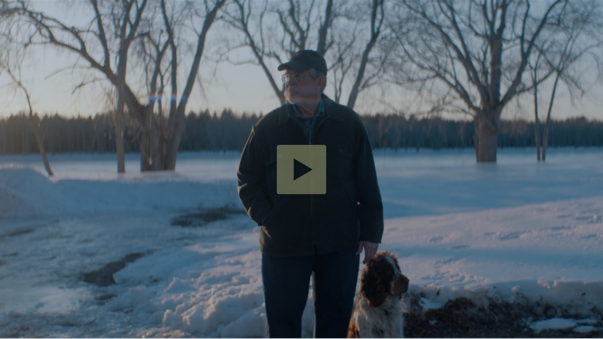 An elderly man wearing a black jacket and cap standing outdoors in a snowy landscape with bare trees in the background, accompanied by a brown and white dog sitting beside him during sunset.