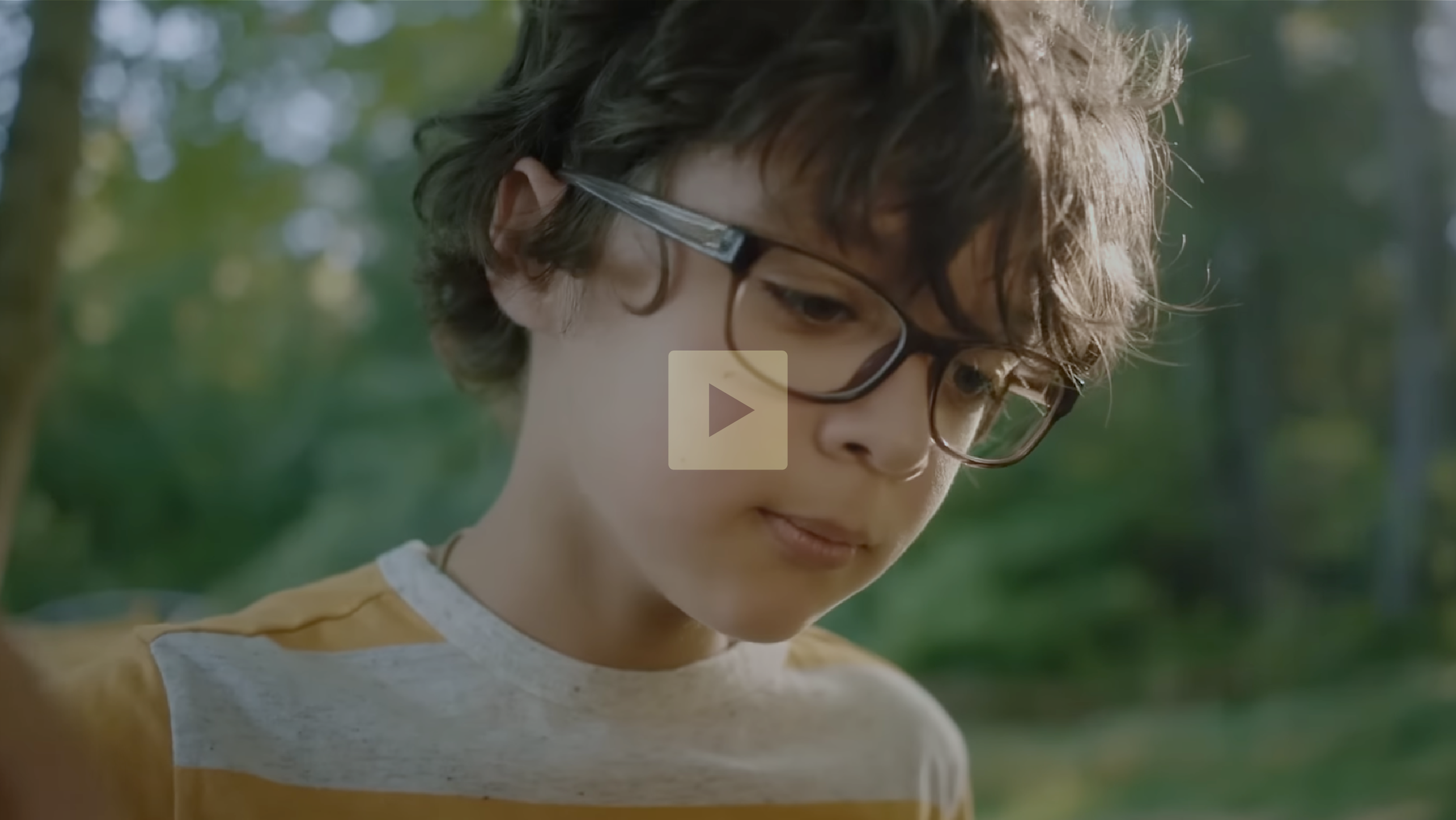 A young boy with curly dark hair, wearing glasses and a yellow and white striped shirt, is outdoors in a forested area during daytime, looking down with a thoughtful expression.