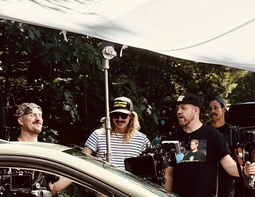Filmmakers and crew filming outdoors under a white canopy, with trees in the background, using professional cameras and equipment.