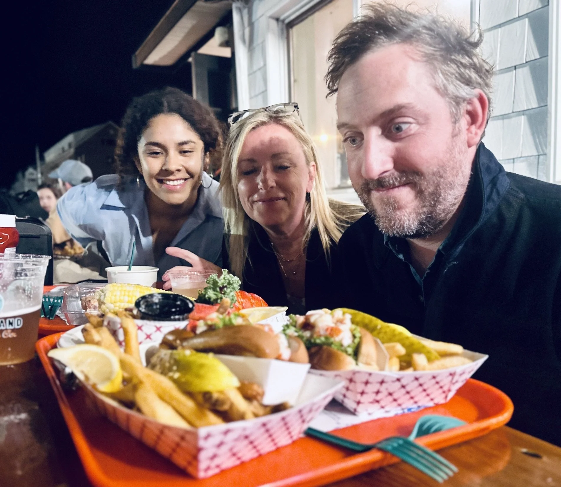 A group of three people smiling at a table with different dishes of food, including fries, tacos, and fried chicken, during a meal at a restaurant.