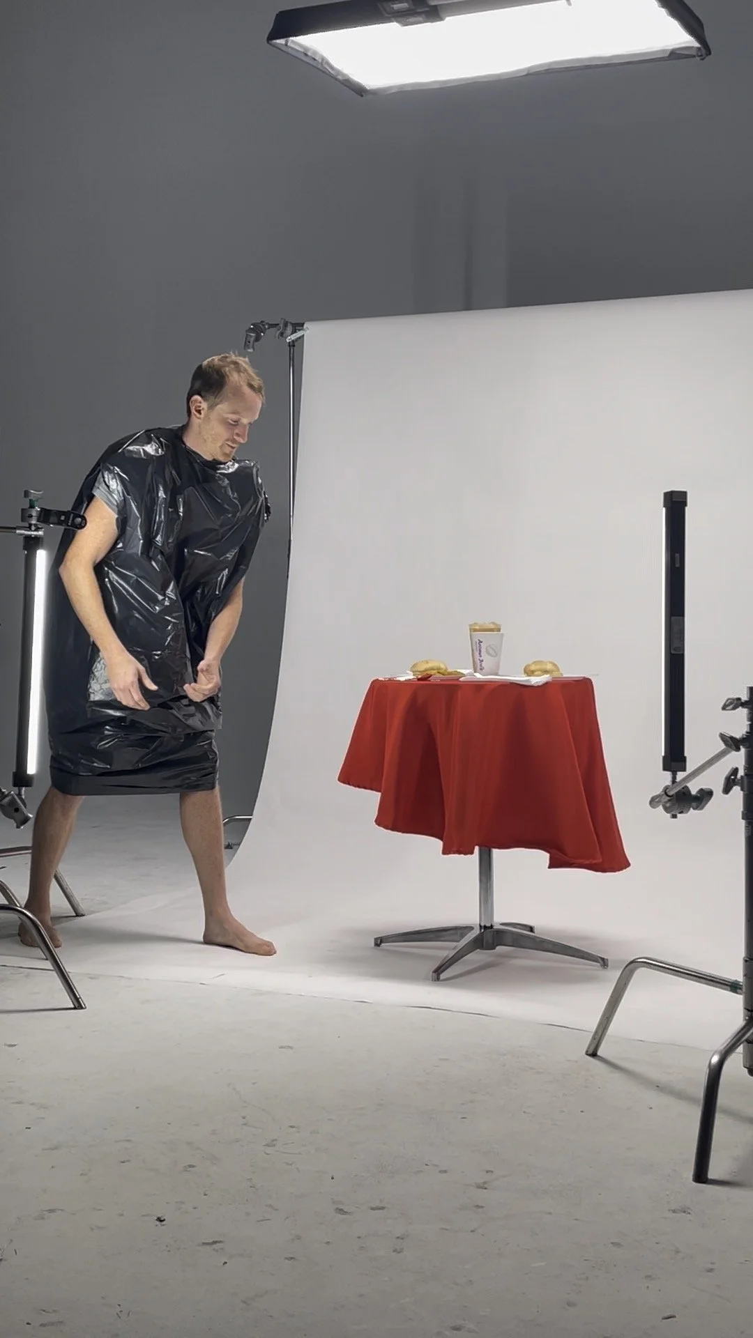 A man in a plastic black apron stands barefoot near a table with a red cloth, holding a sandwich and a beverage cup, in a photography studio with a white backdrop and studio lights.