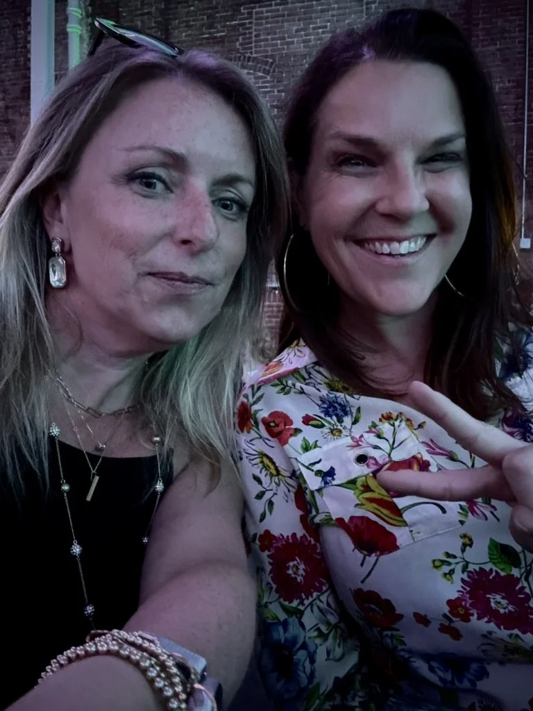 Two women smiling and taking a selfie outdoors at night. One woman has blonde hair, is wearing earrings, necklaces, and a black top. The other woman has dark hair, is making a peace sign, and wearing a colorful floral top. There is a brick wall in th