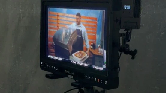 A video monitor shows a man preparing food in a kitchen or cooking studio with orange paneling in the background.