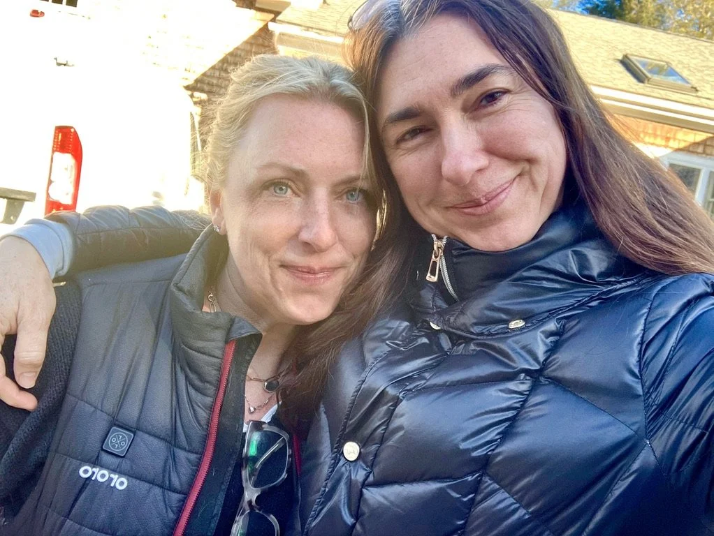Two women taking a selfie outdoors, smiling, with a house and a garage door visible in the background. One woman has blonde hair and blue eyes, wearing a black vest, and the other has brown hair, wearing a dark blue puffer jacket.