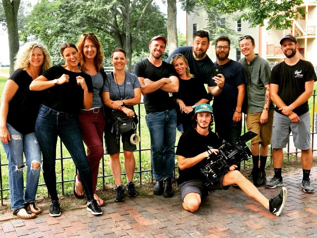 Group of eleven people smiling and posing outdoors on a brick path, with trees and residential buildings in the background, some with cameras and equipment.