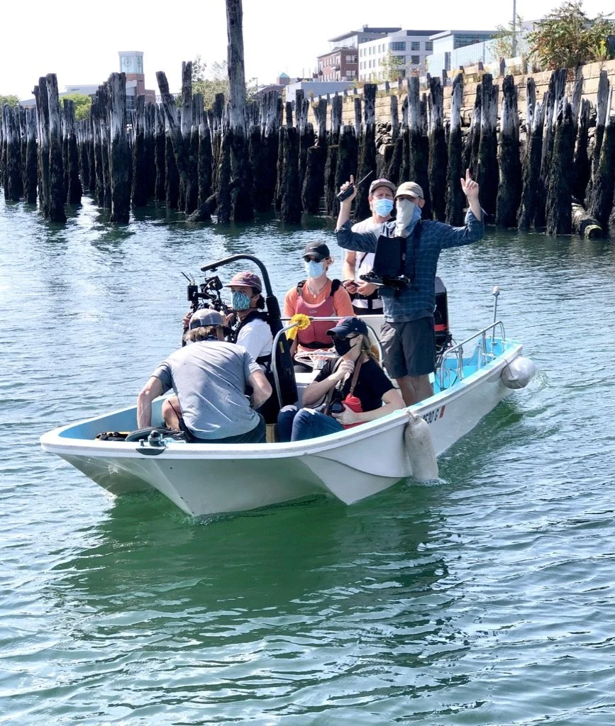 A group of people on a small boat, some with masks, with two standing individuals holding equipment, near a pier with wooden posts and a cityscape in the background.
