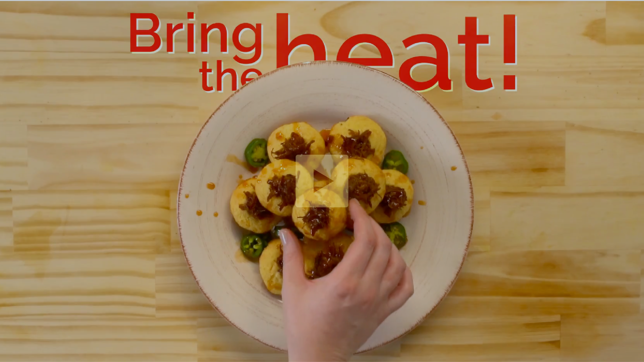A top-down view of a bowl filled with bite-sized potato balls topped with caramelized onions and surrounded by sliced green jalapeños, with a hand stretching over to pick one up. Red text reads "Bring the heat!"
