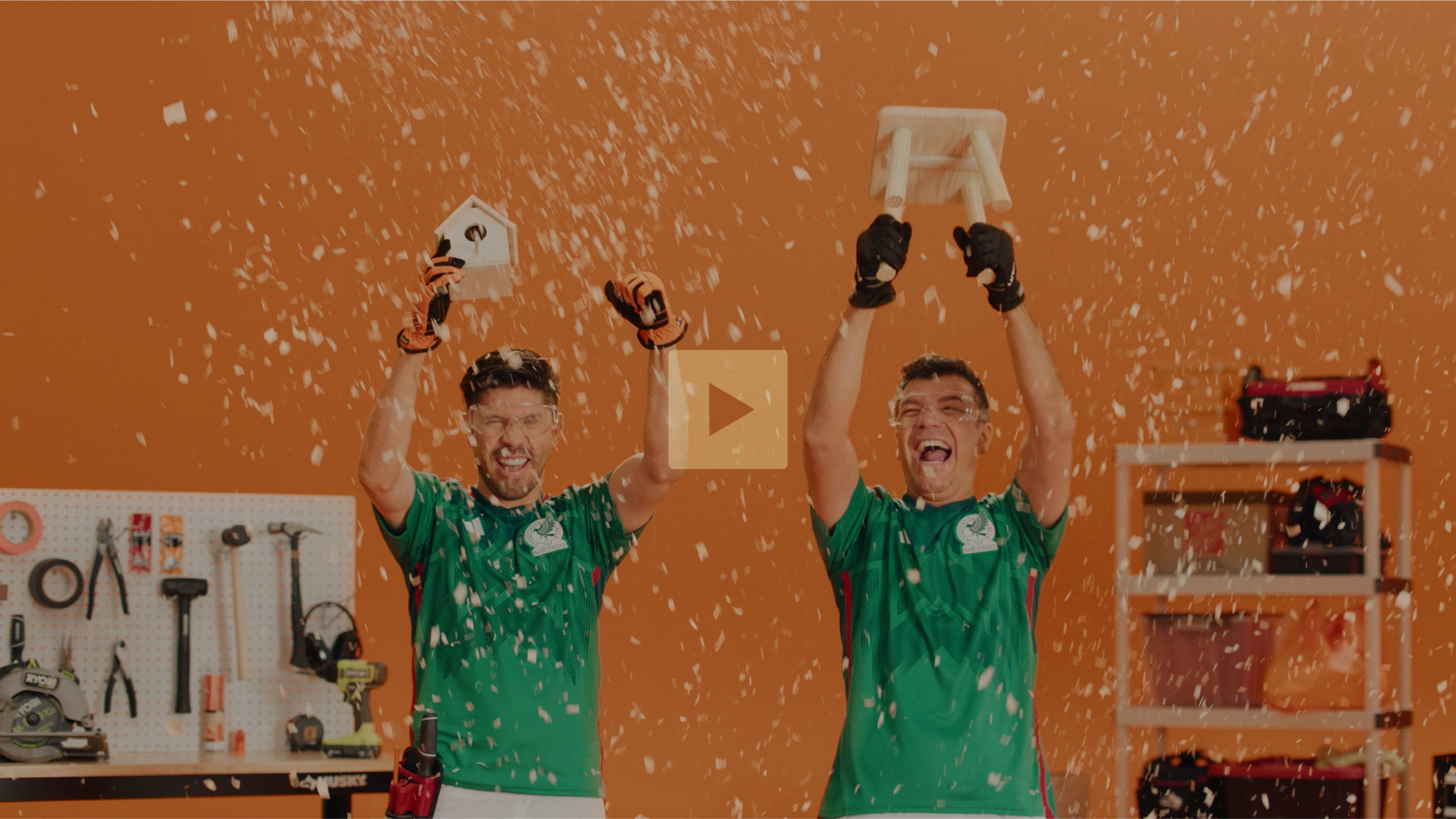 Two men wearing green sports jerseys celebrating and holding trophies in a workshop, with confetti falling around them.