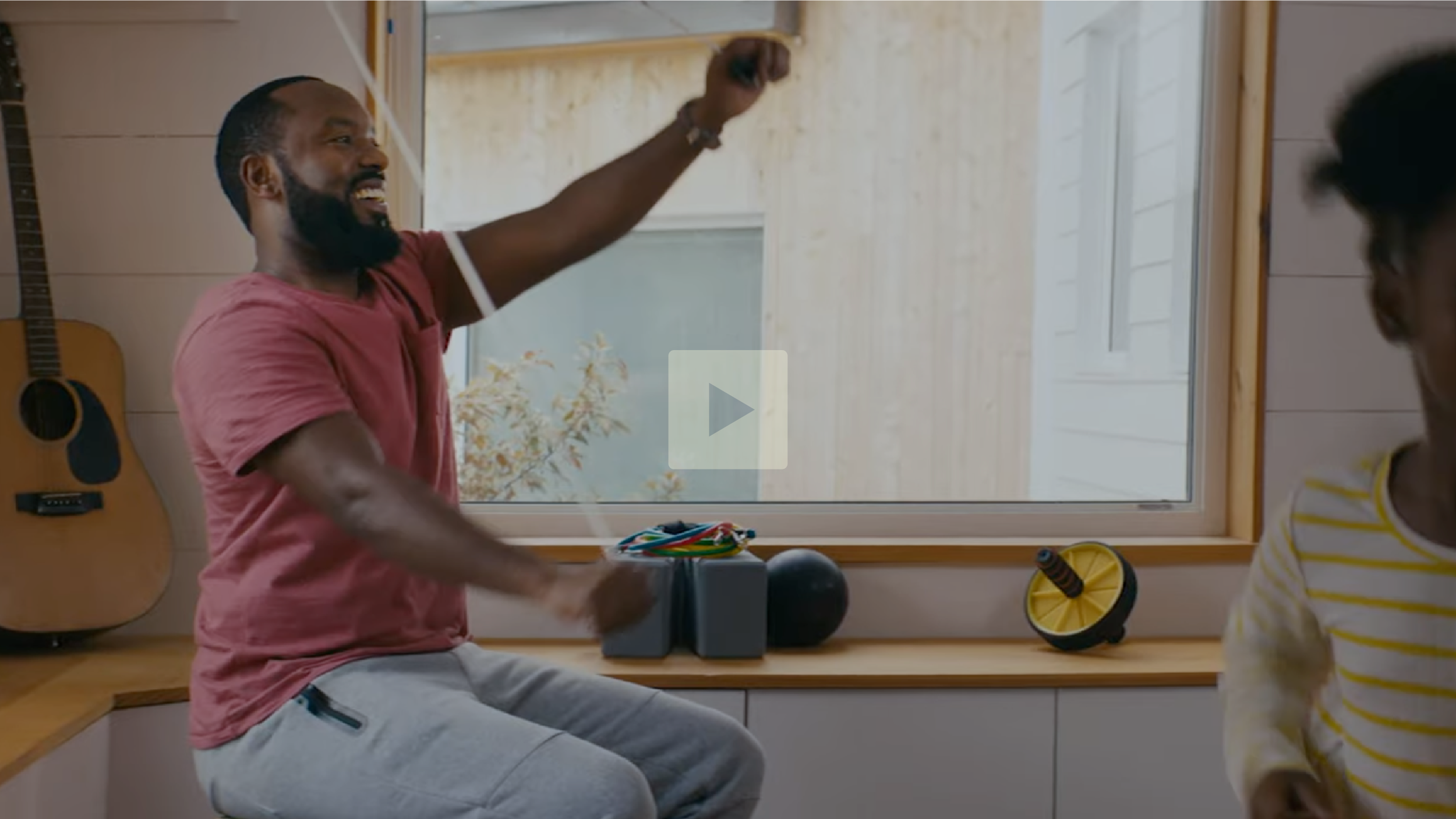 A man with a beard smiling and holding colorful juggling clubs, sitting on a kitchen counter next to a window. There is a guitar hanging on the wall and various juggling props on the counter.