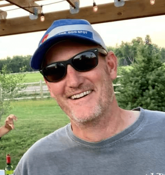 A smiling man wearing sunglasses and a blue cap outdoors during the daytime, with green trees and a wooden structure with string lights in the background.