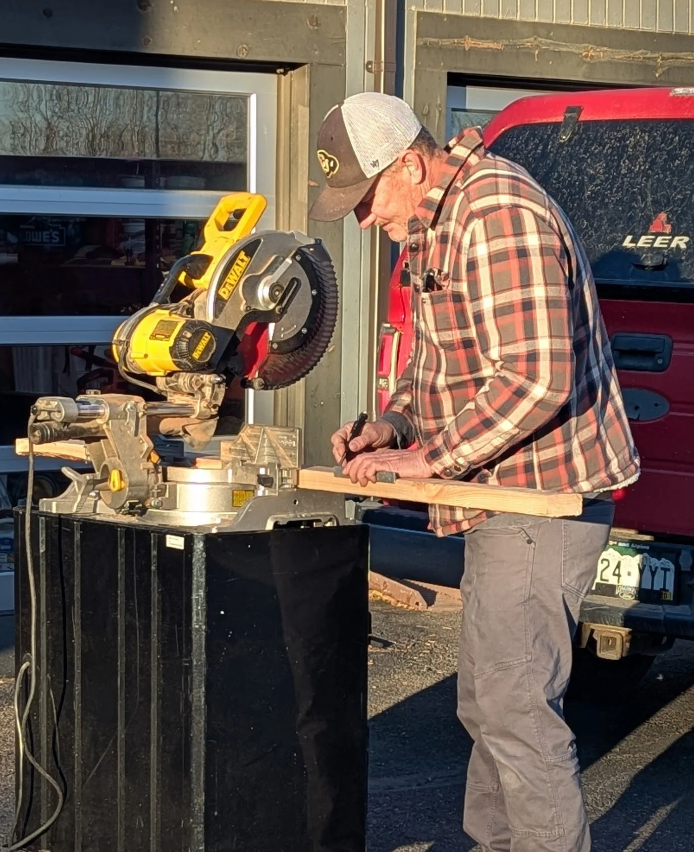 A man wearing a plaid shirt, baseball cap, and gloves is using a miter saw to cut a piece of wood outdoors in front of a garage.