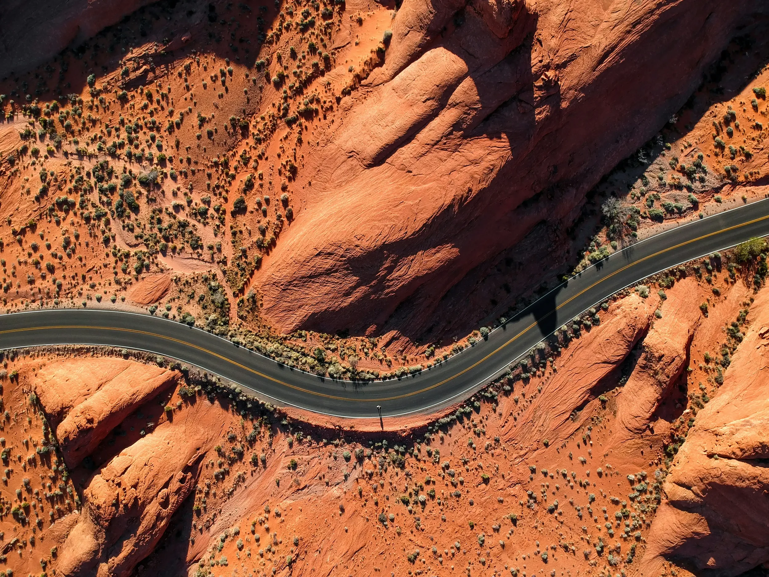 An aerial top-down view of a winding asphalt highway cutting through dramatic red rock desert formations, representing the Epic Road Trips driving itineraries on Swallow's Notes.
