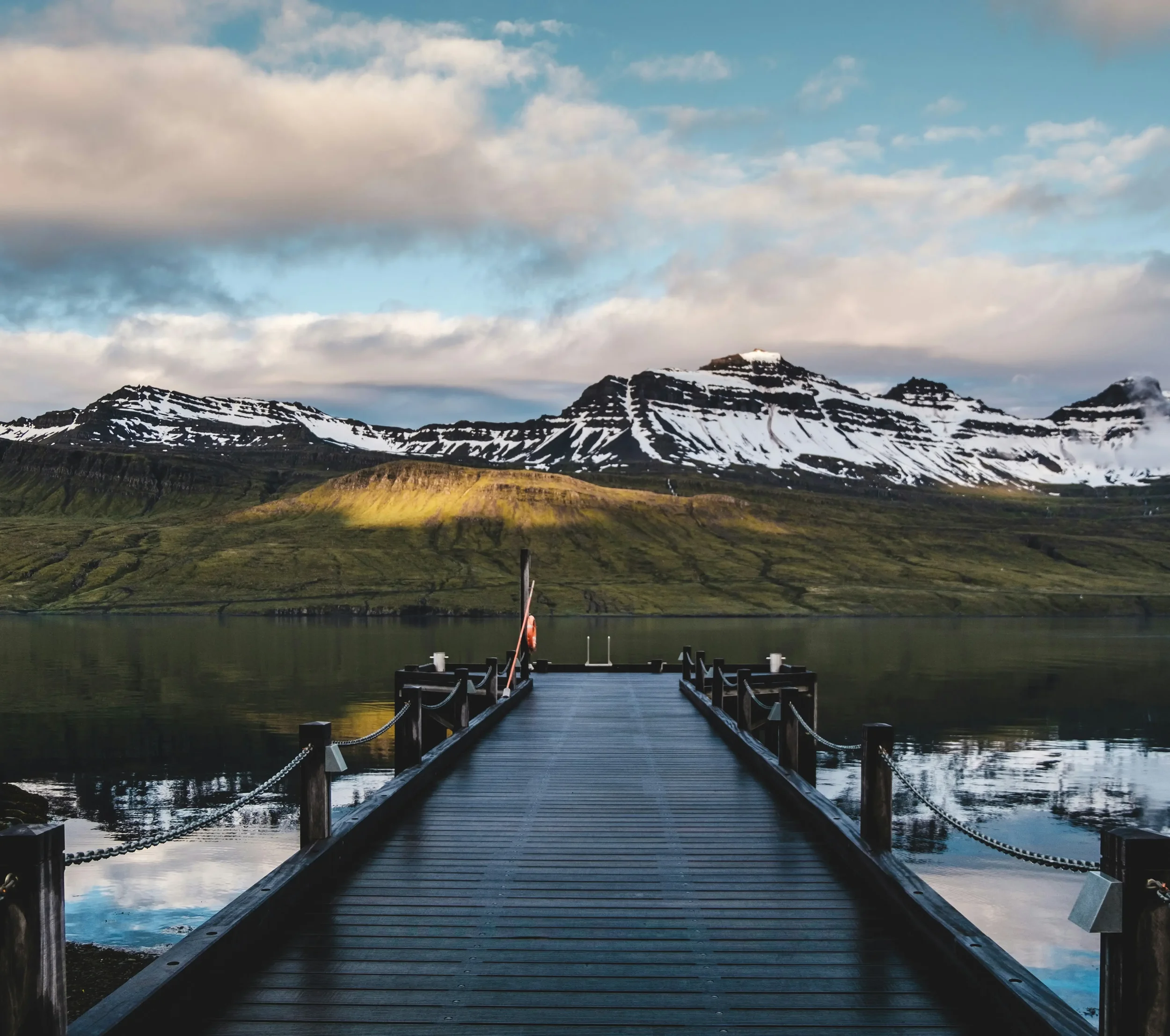 Panoramic view of steep, snow-capped mountains and calm blue water in a narrow bay of the East Fjords, Iceland, with a quiet coastal road in the foreground.