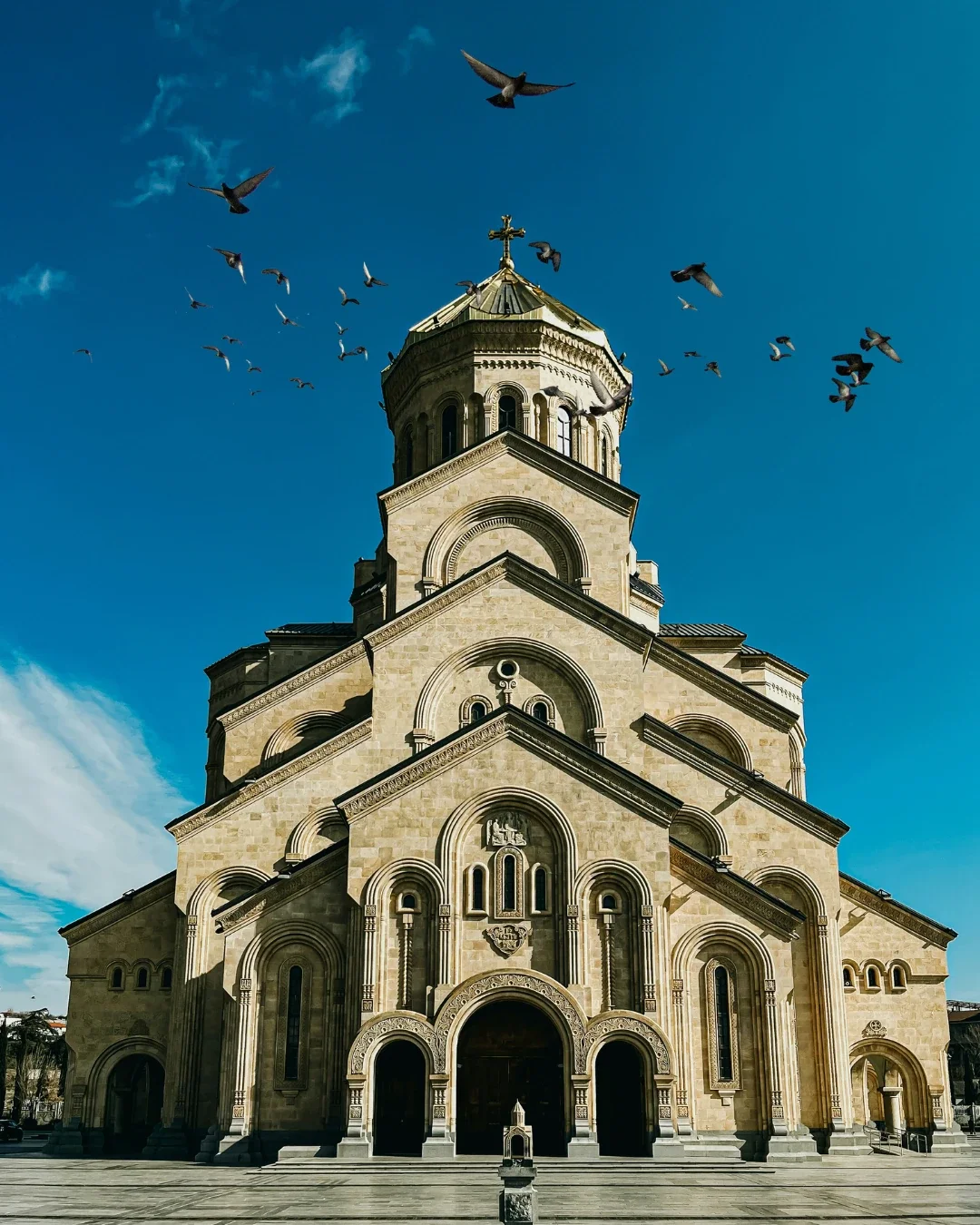 The Holy Trinity Cathedral of Tbilisi (Sameba) in Georgia, featuring its massive stone facade and golden dome with pigeons flying in a clear blue sky.