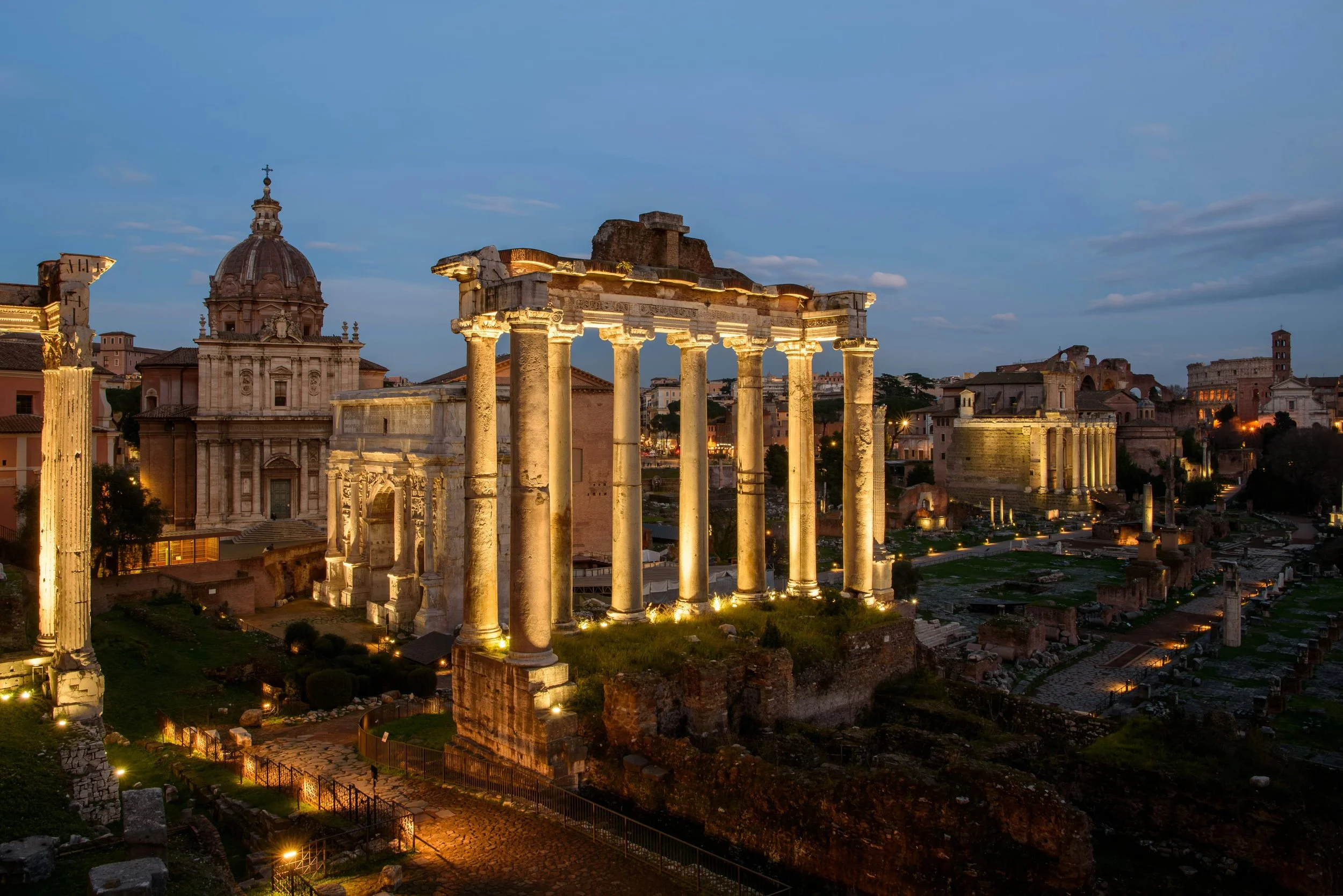 The ancient ruins of the Roman Forum illuminated at dusk with the Temple of Saturn in the foreground.