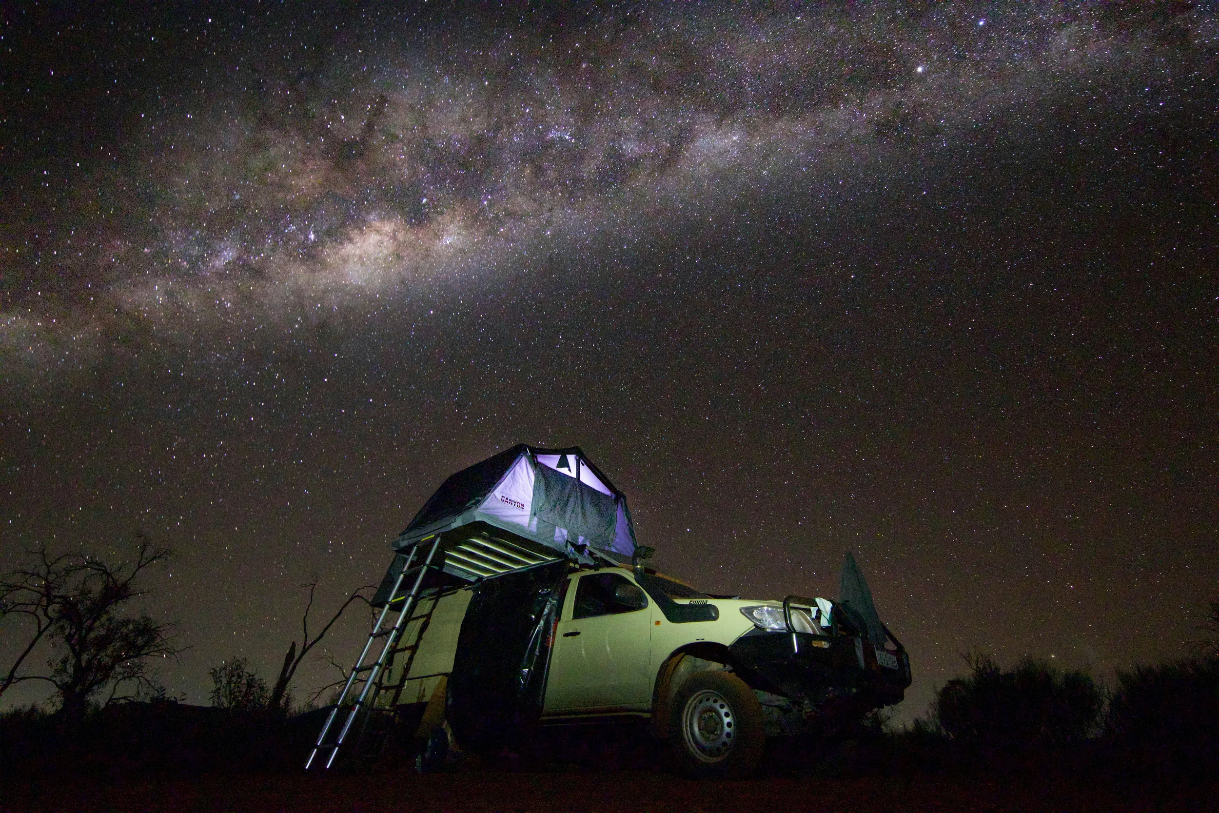 A rugged 4WD expedition vehicle with an open roof-top tent parked in the Australian outback under a spectacular starry night sky and the Milky Way.
