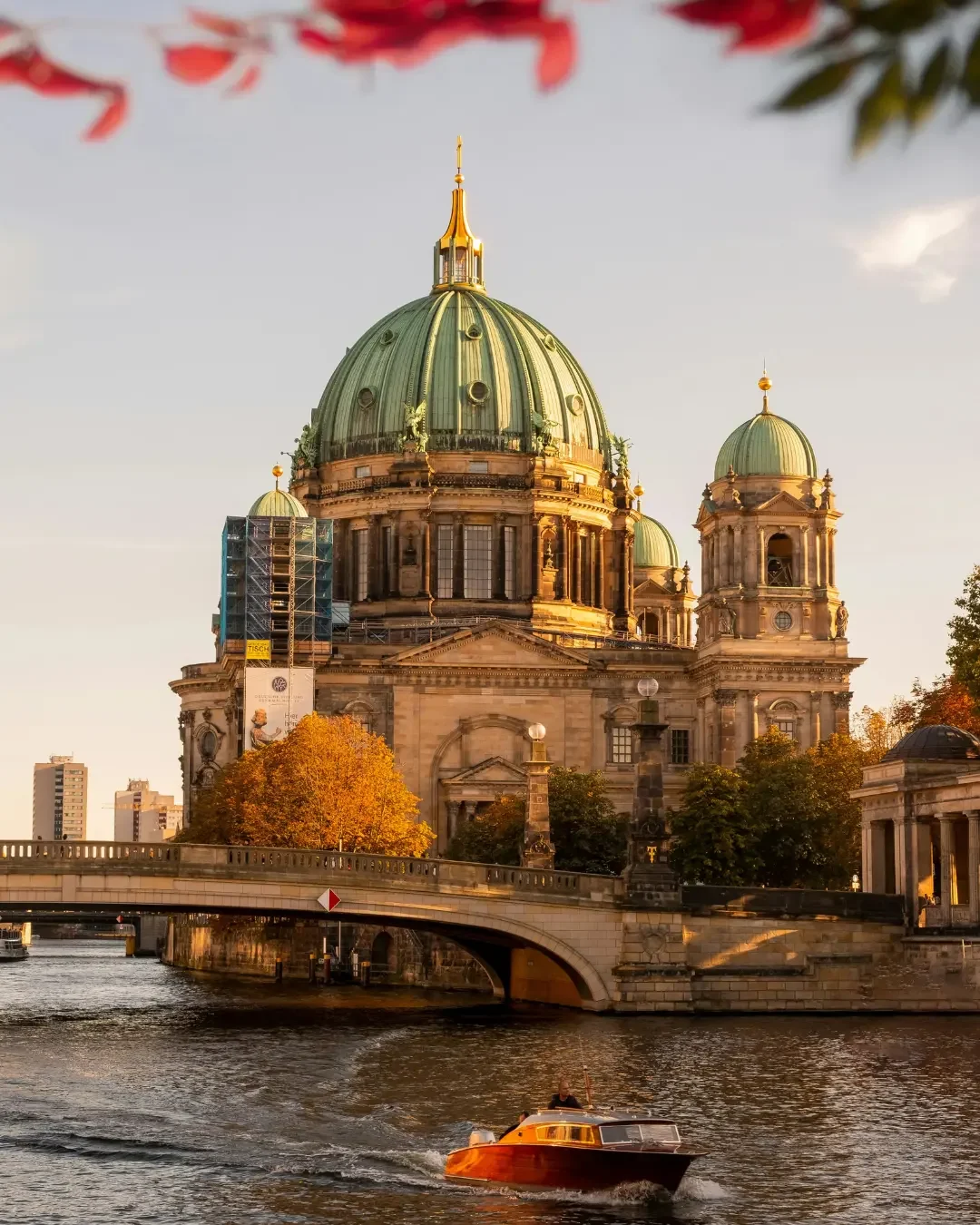 Baroque-style cathedral with a large central green dome, smaller surrounding domes, situated along a river with a boat passing by, in warm sunset light.