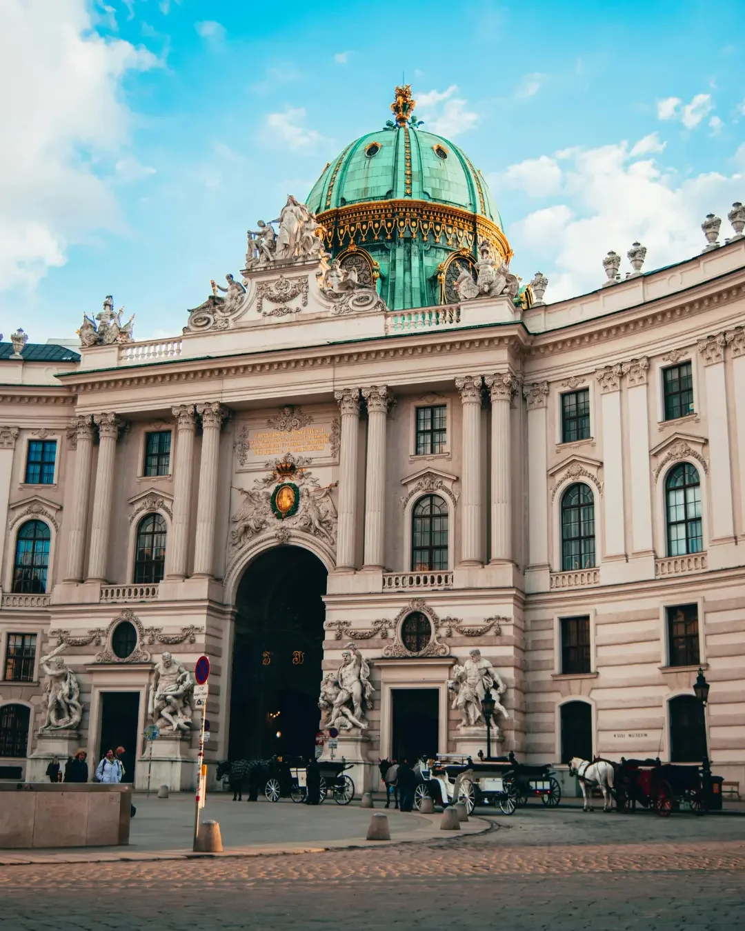 Baroque palace with a green copper dome, white stone sculptures, and columns, featuring horse-drawn carriages in front under a blue sky.