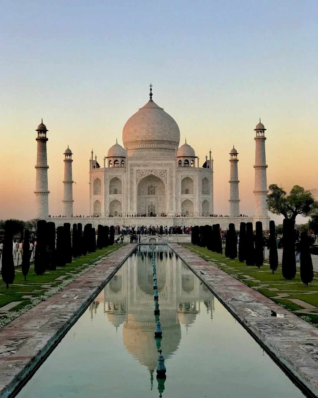 The Taj Mahal during sunset with visitors around, a water reflecting pool in front, and trees on the sides.