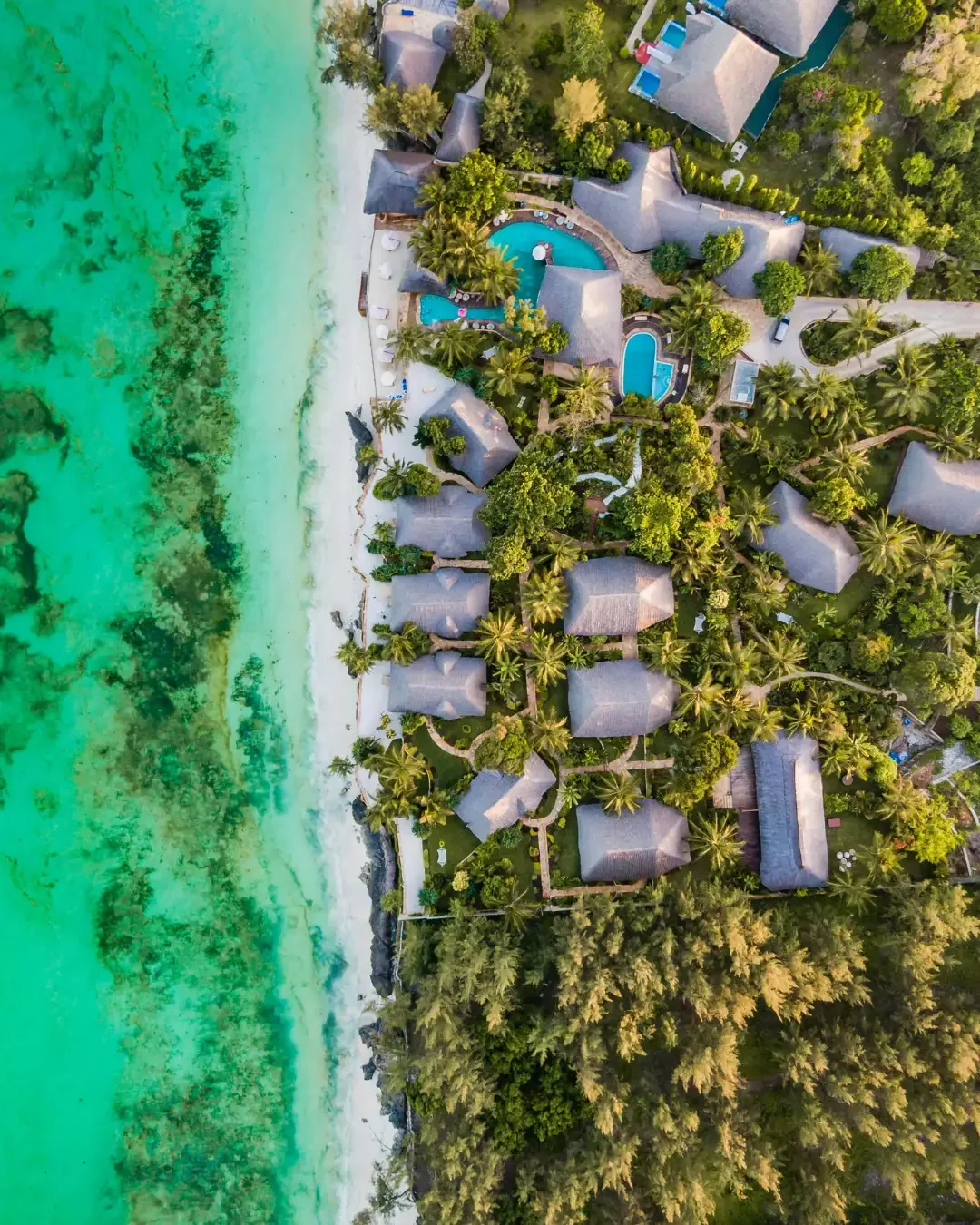 An aerial top-down view of a luxury beachfront resort in Zanzibar, Tanzania, featuring thatched-roof villas, turquoise ocean water, and lush tropical palm trees.